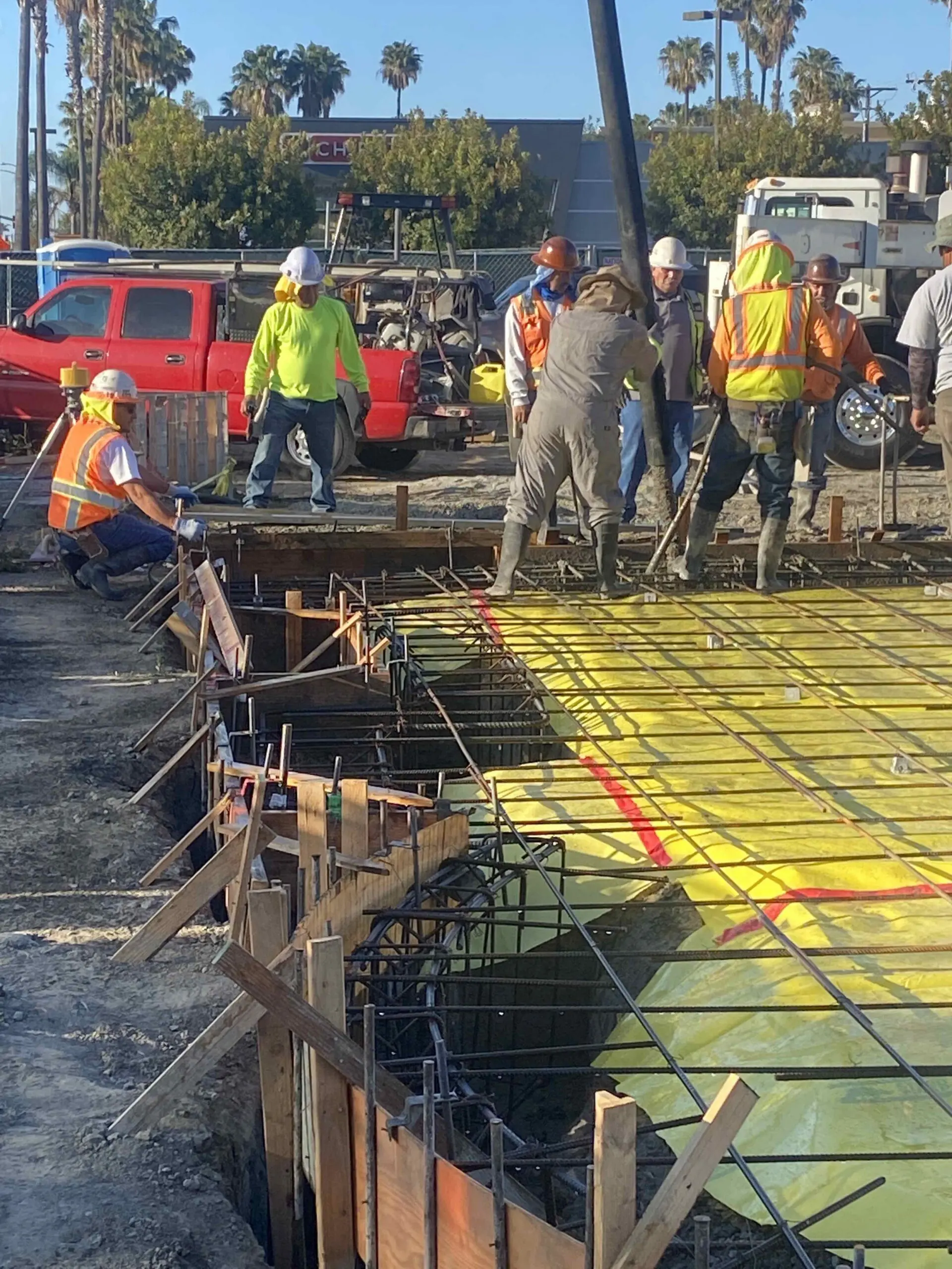 Group of men wearing helmets and working on a construction site in, are vehicles on the road, trees, buildings, poles