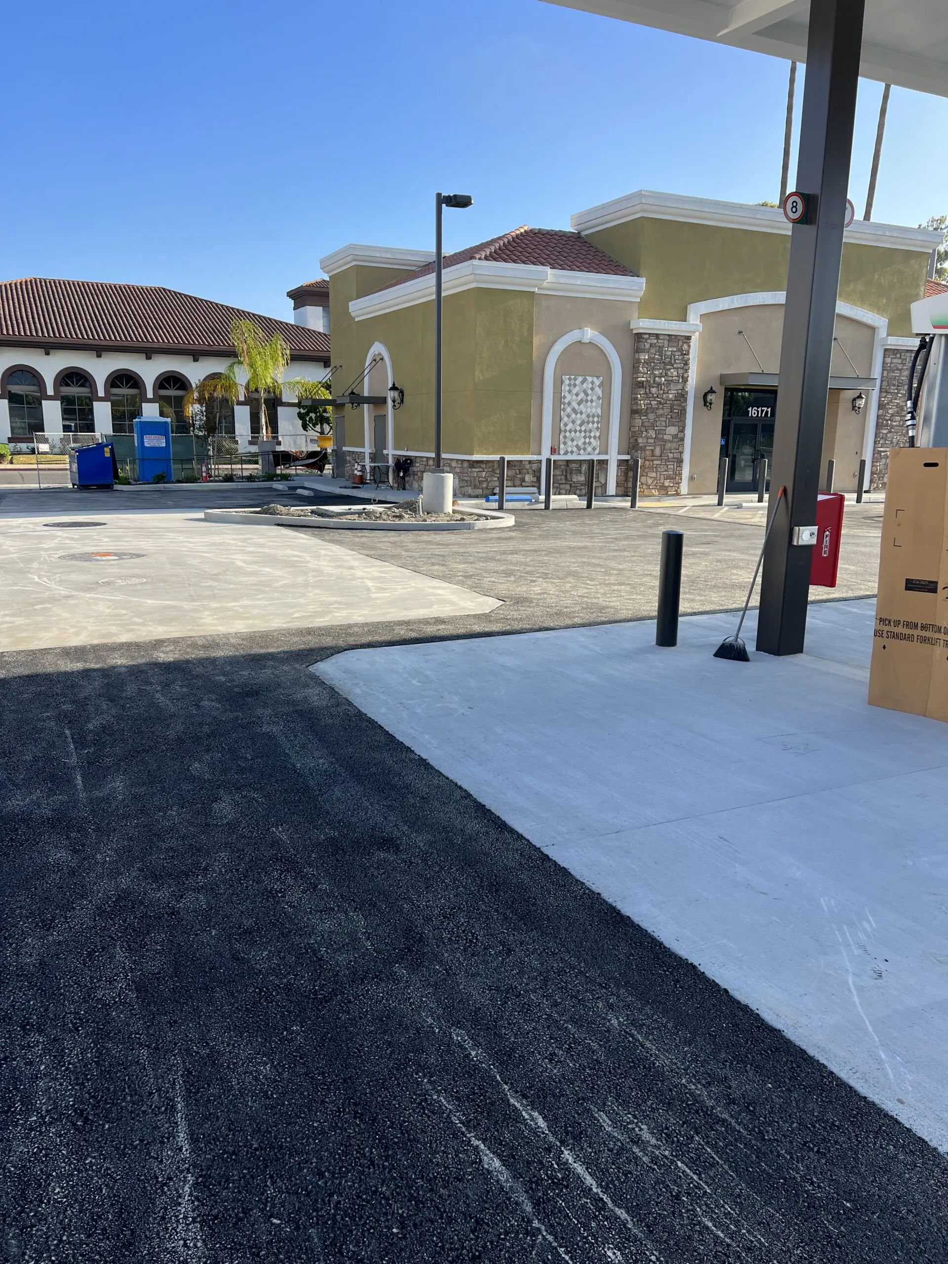 Parking lot with a gas station in the background, surrounded by buildings, trees, poles, lights, boards, dustbins