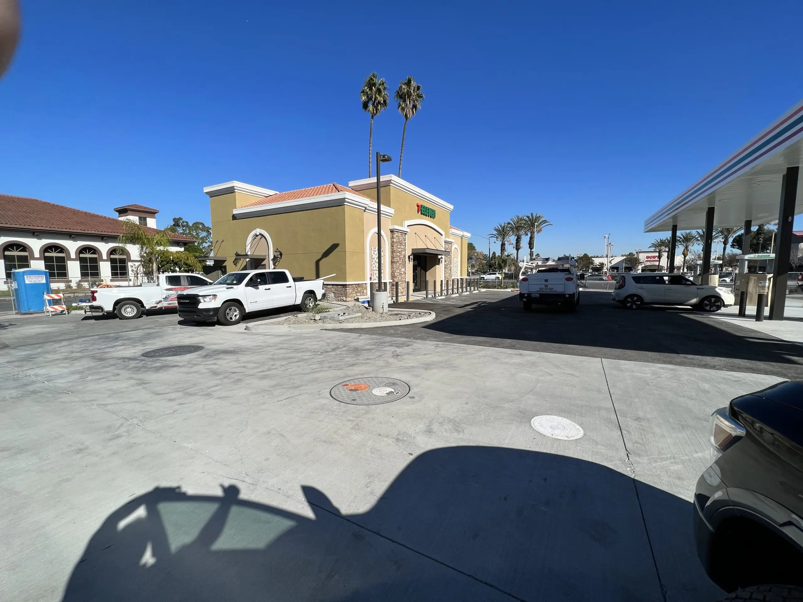 Parking lot with cars parked in front of a building, surrounded by trees, poles, and other objects