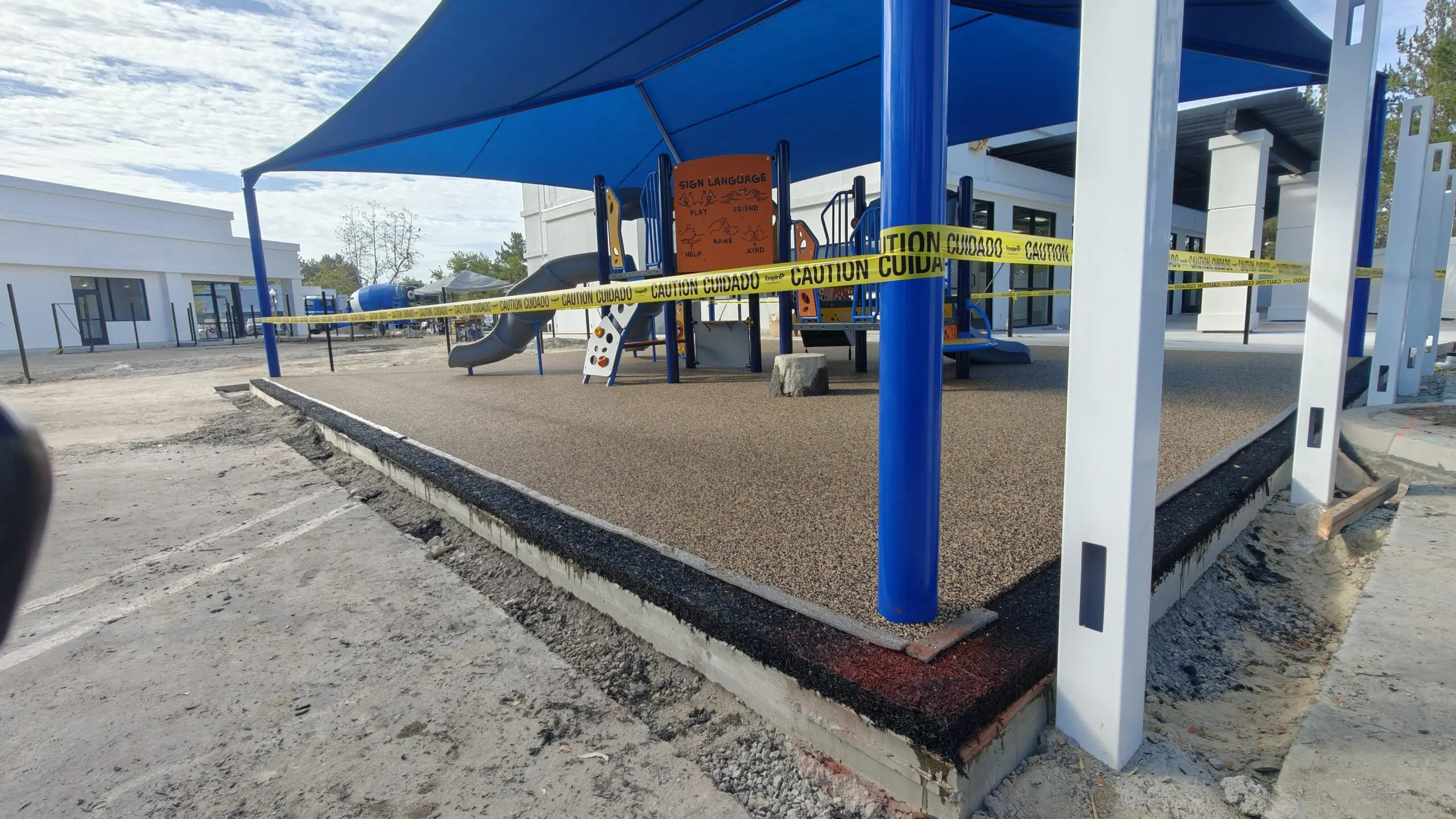 Playground with a blue canopy over it, surrounded by buildings, trees, poles, and a yellow caution tape