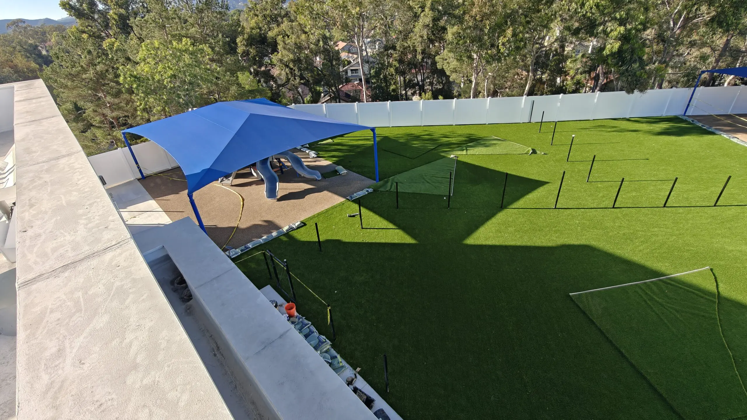 Backyard with artificial grass and a blue shade sail, surrounded by a wall and trees