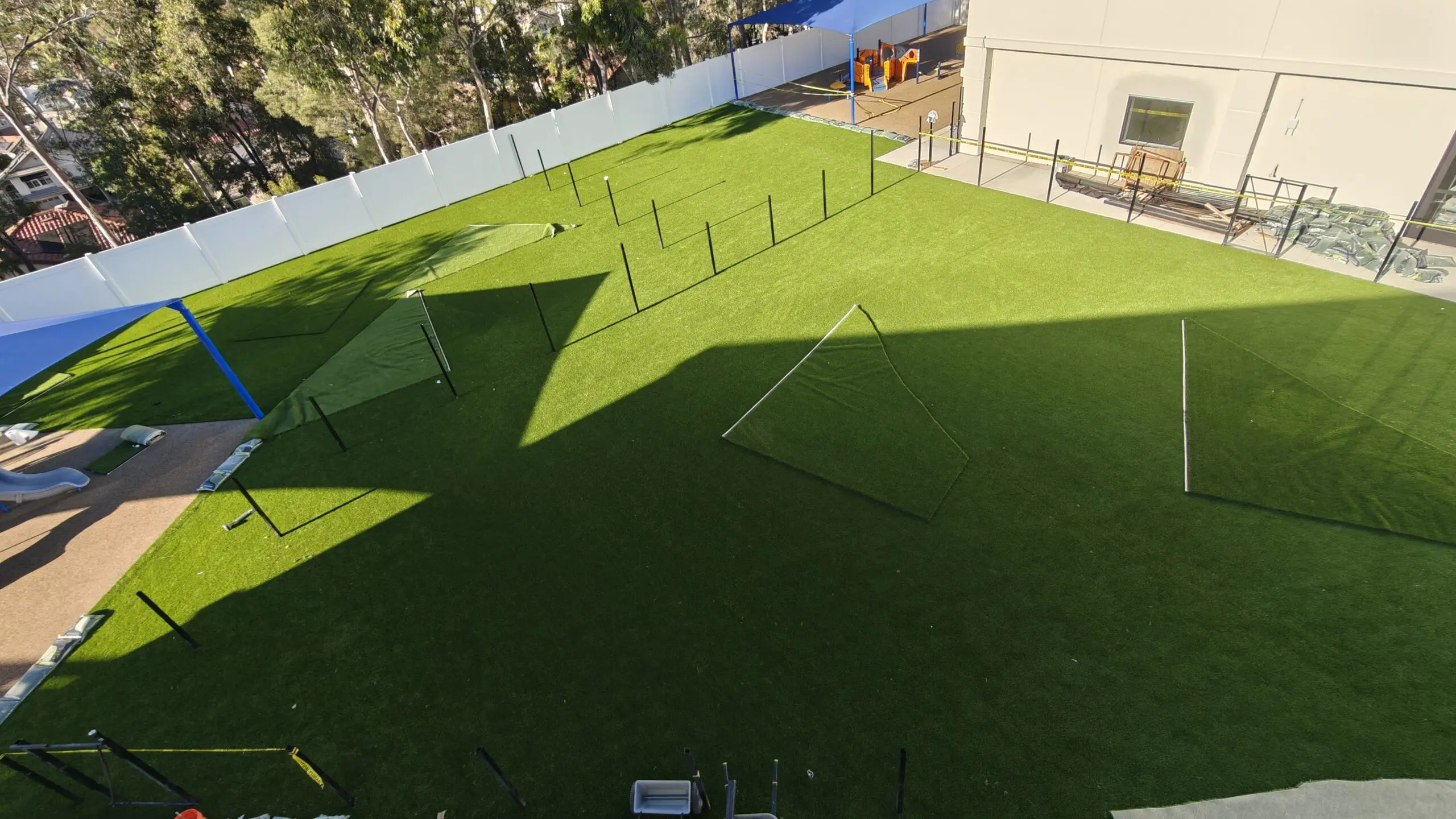 Aerial view of a playground with artificial grass, a fence, poles, a tent, a slide, a building with windows