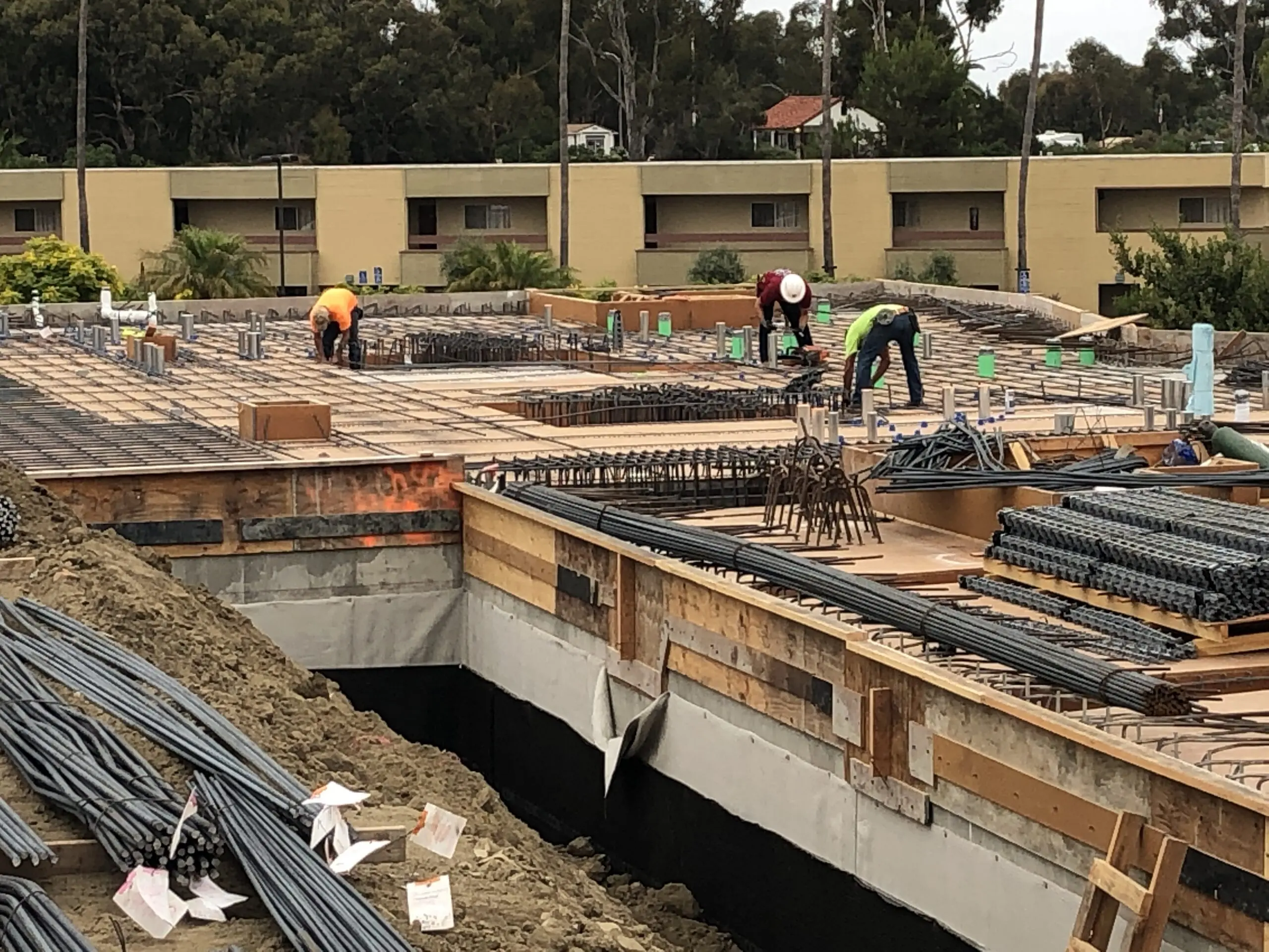 Group of men working on a construction site in front of a building, surrounded by iron rods, wooden objects