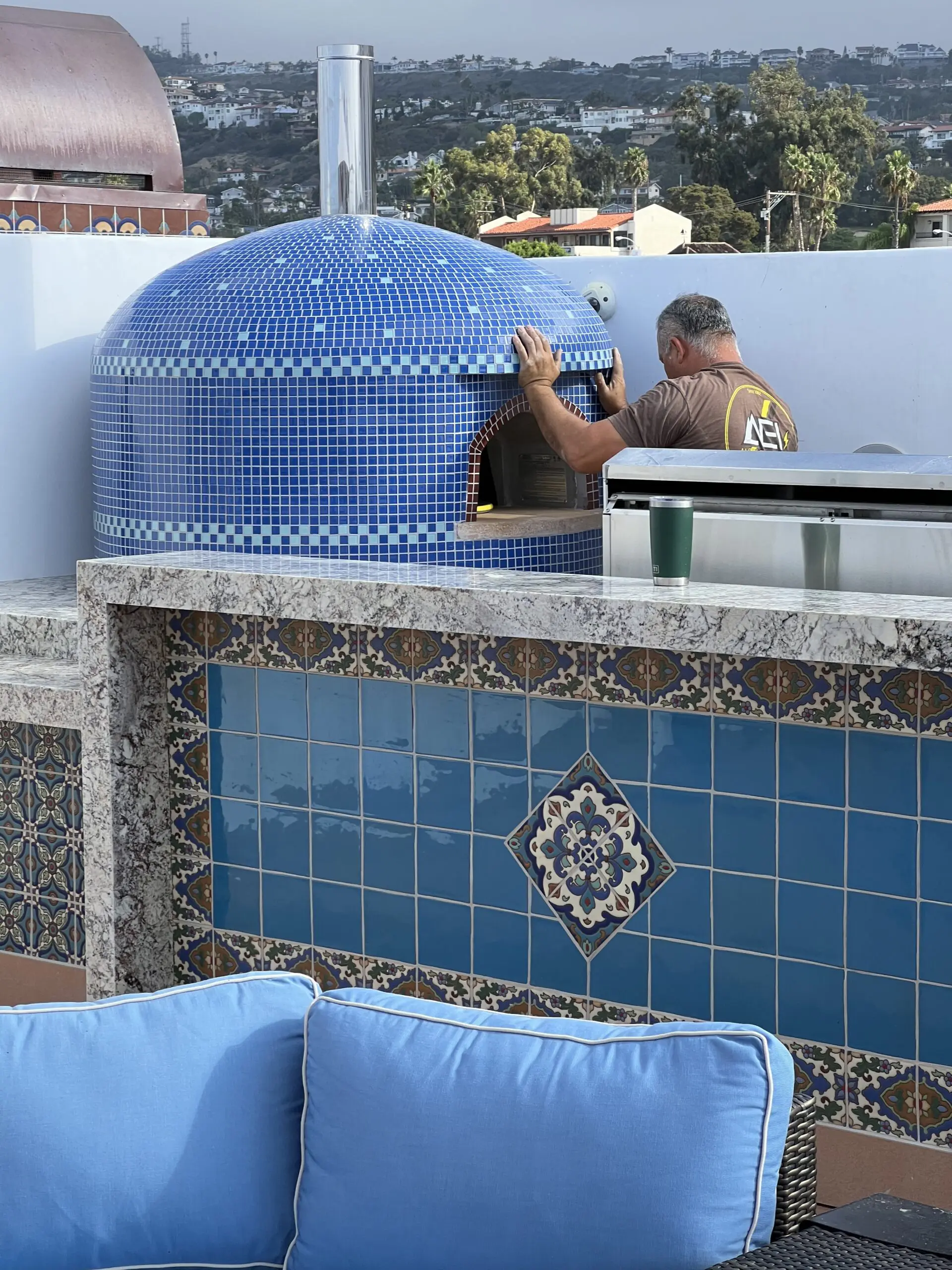 Man standing on top of a roof next to a blue couch with pillows, a coffee cup, and a lamp