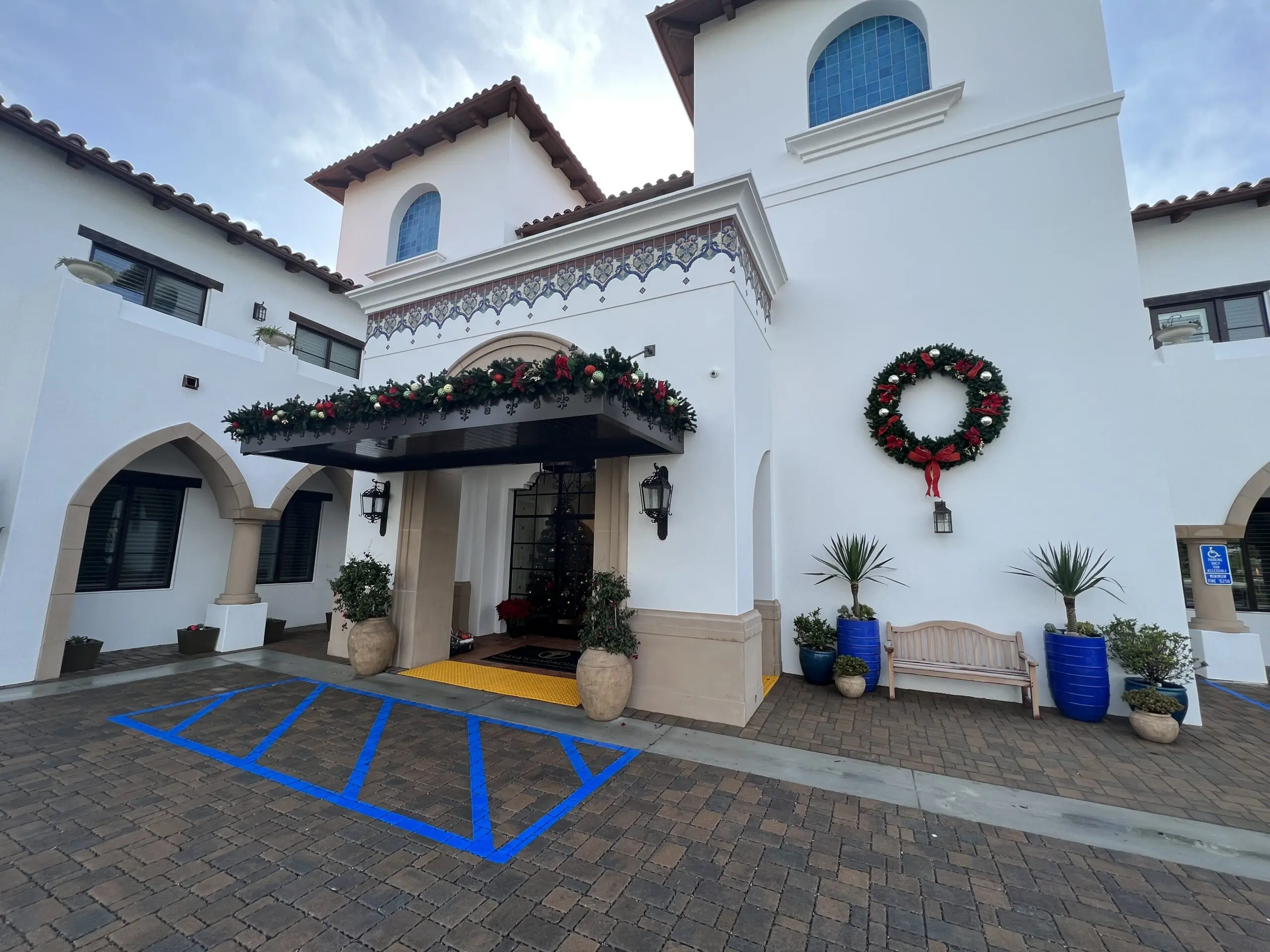 White building with a wreath on the front of it, surrounded by houseplants, a bench, and a lamp