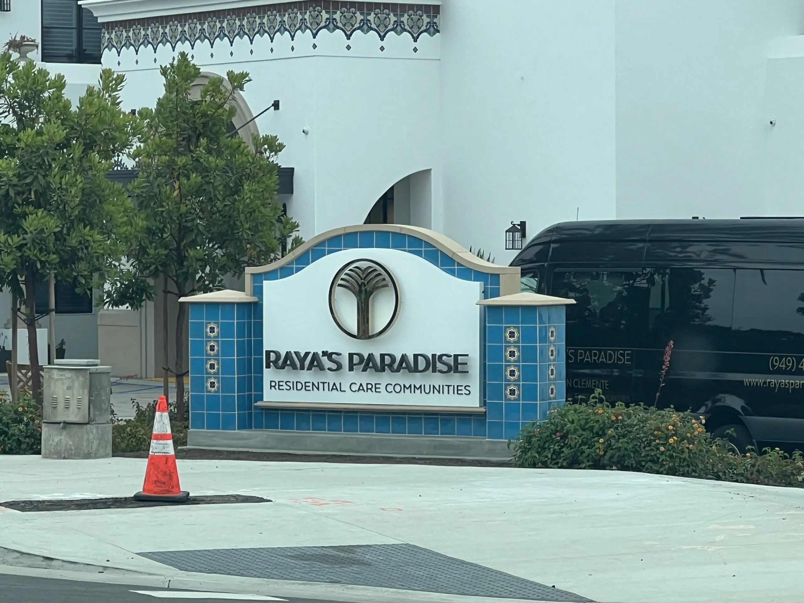 Building with a sign that reads "Rayas Paradise Residential Care Communities" in front of it, surrounded by trees, plants