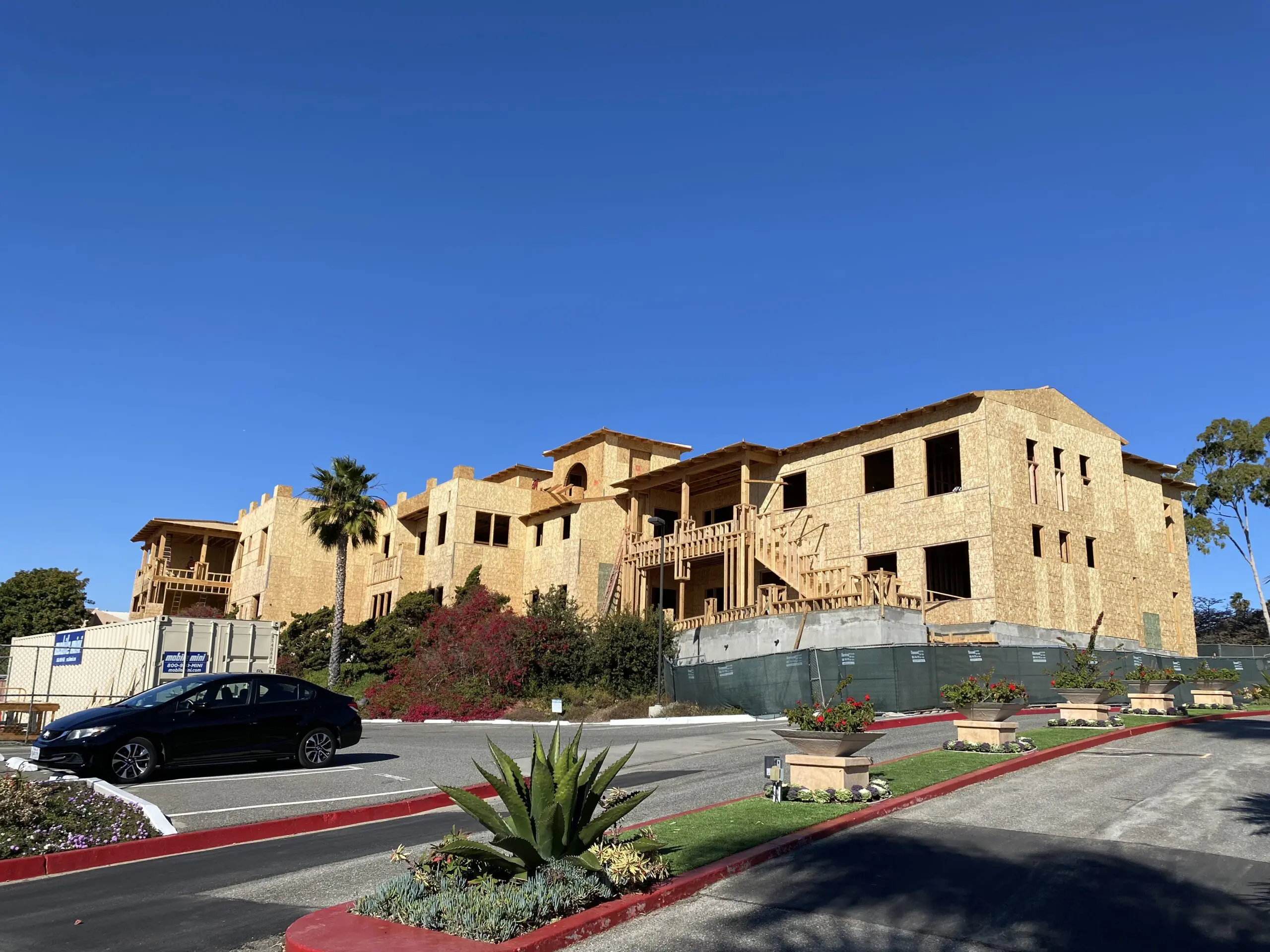 Large building under construction in the middle of a street, surrounded by trees, plants, and grass