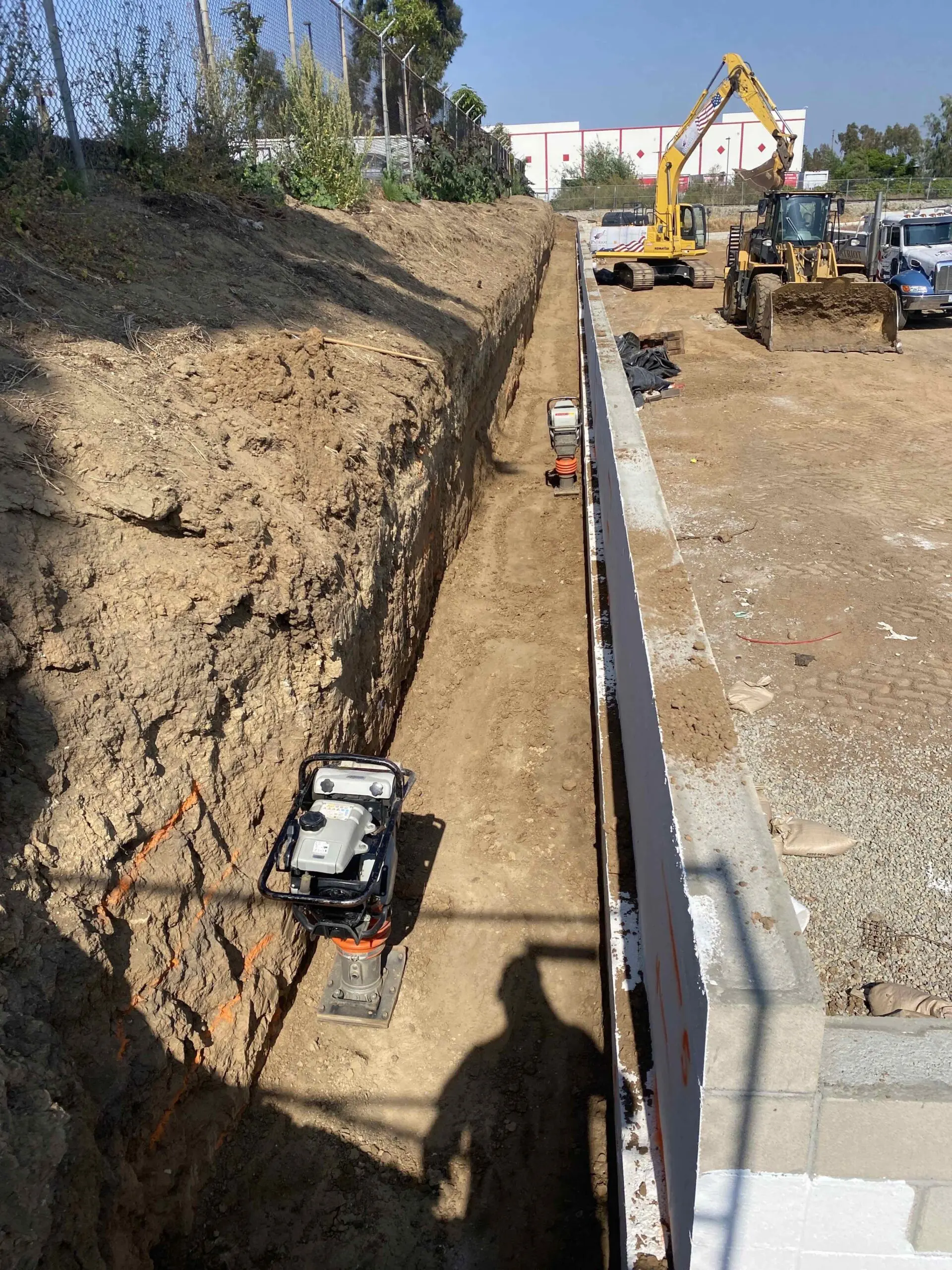 Construction site with a large pile of dirt next to a fence, surrounded by vehicles, trees, plants