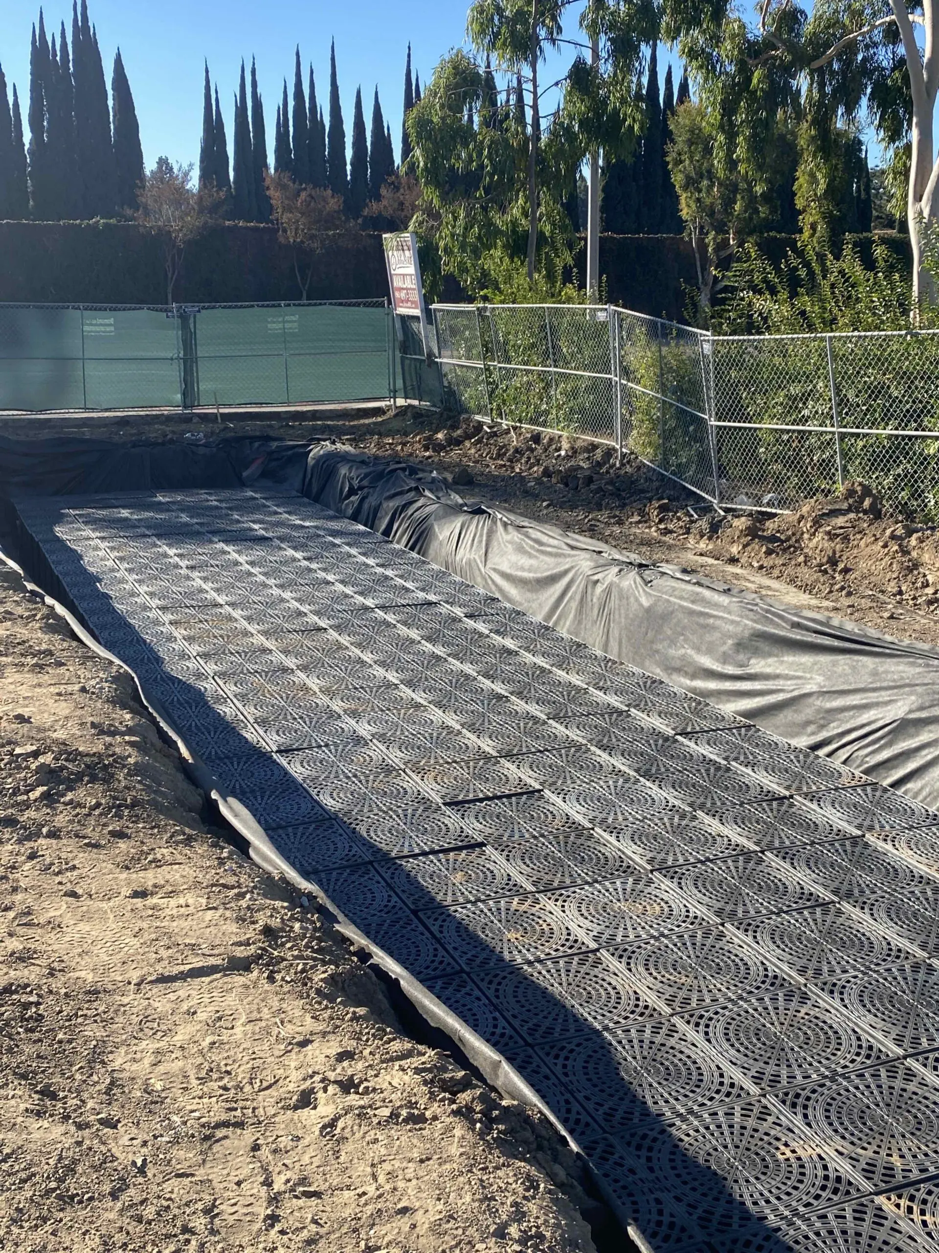 Construction site with a large pile of dirt next to a fence, surrounded by trees and a clear blue sky