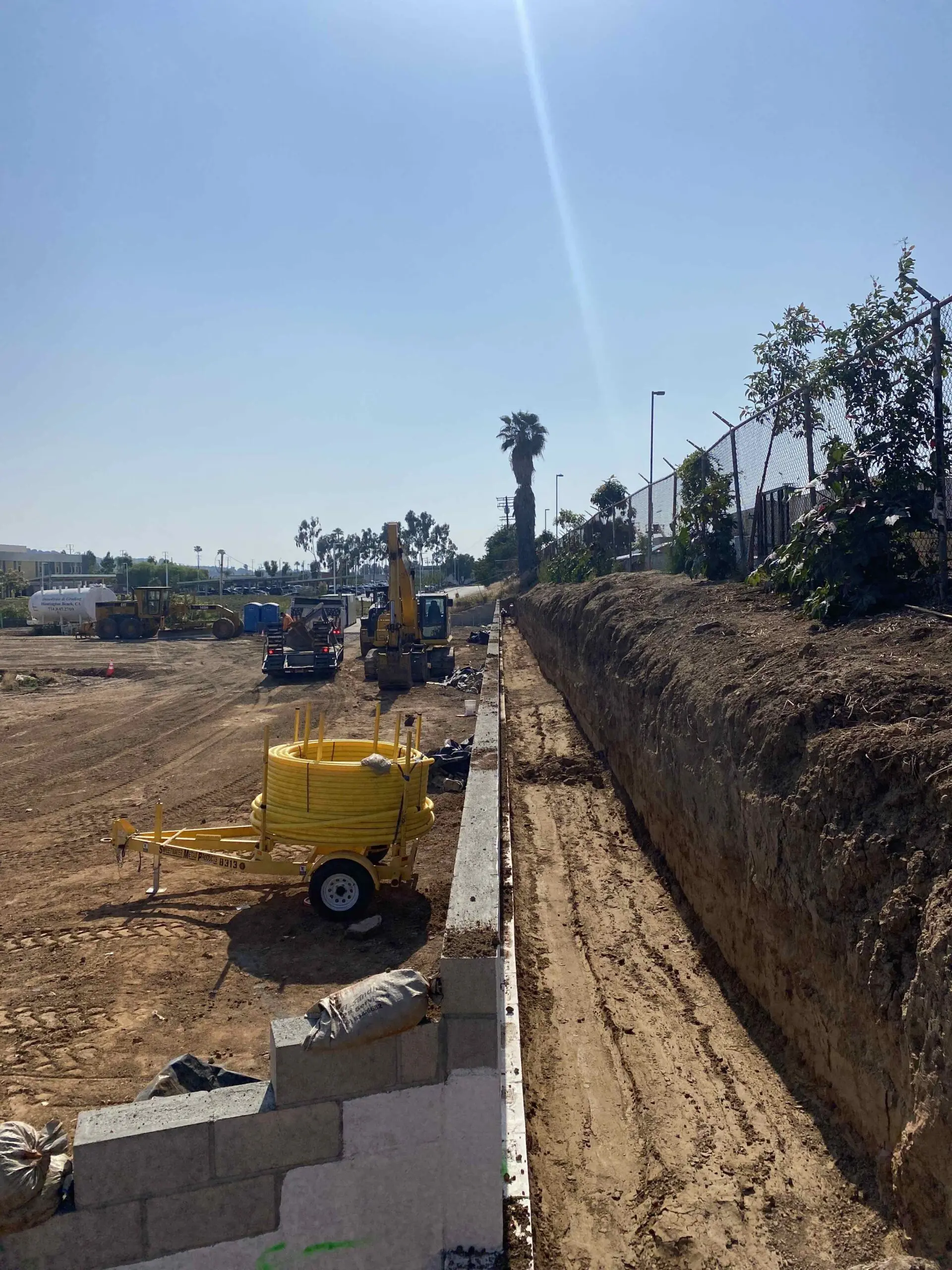 Construction site with a large pile of dirt next to a fence, surrounded by vehicles, trees, poles, and a wall