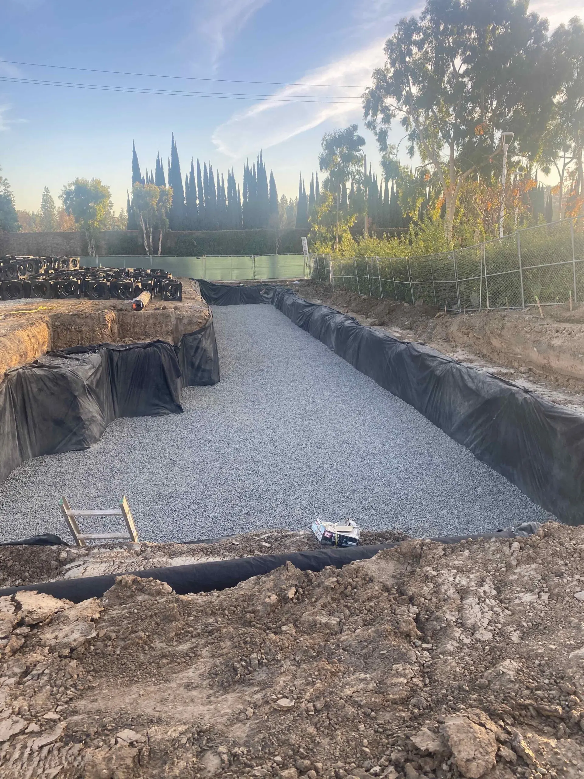 Construction site with a gravel driveway in the middle of it, surrounded by a fence and trees
