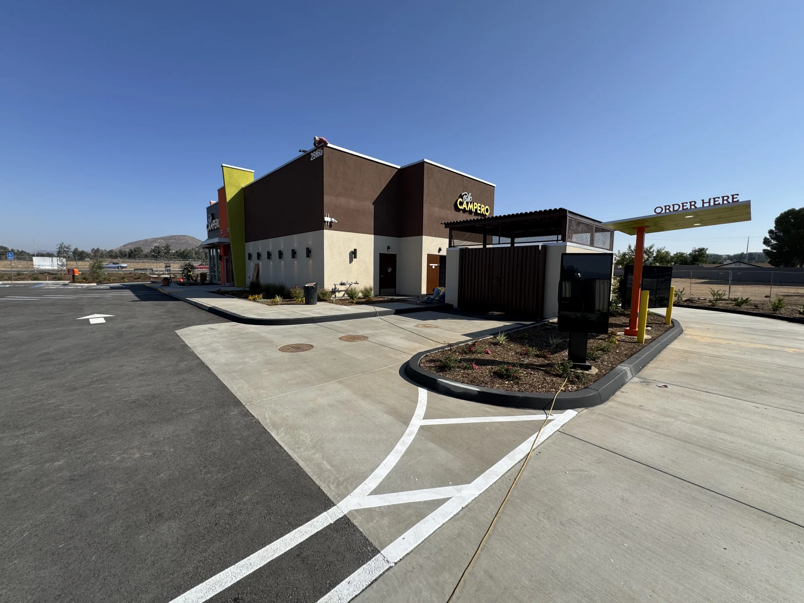 Building with a parking lot in front of it, surrounded by plants, poles, a fence, a group of trees, hills