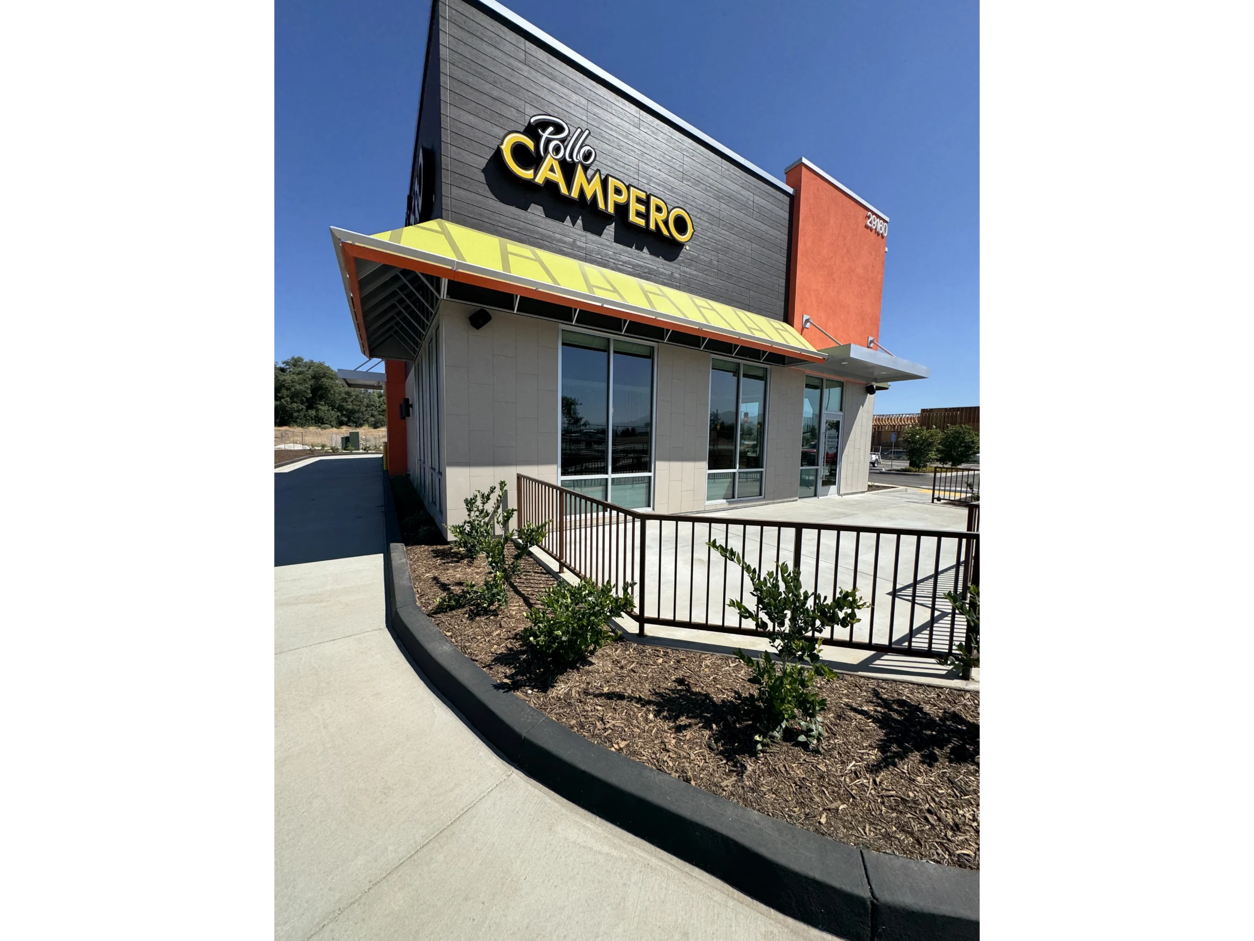 Building with a sign that reads "Pollo Campero" in front of it, surrounded by plants, a metal fence, a pathway