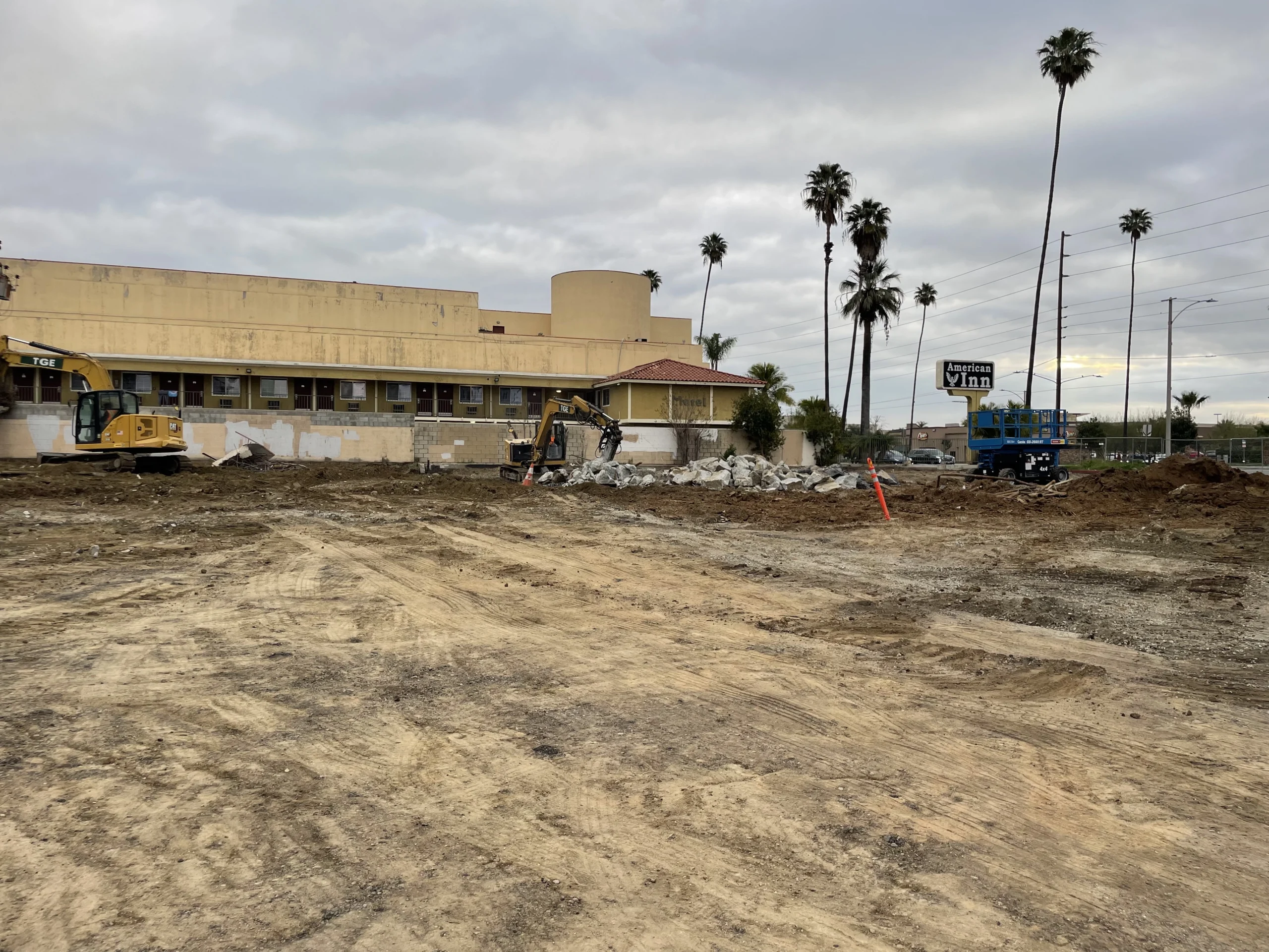 Construction site with a large building in the background, surrounded by cranes, stones, trees, poles, wires, and a board