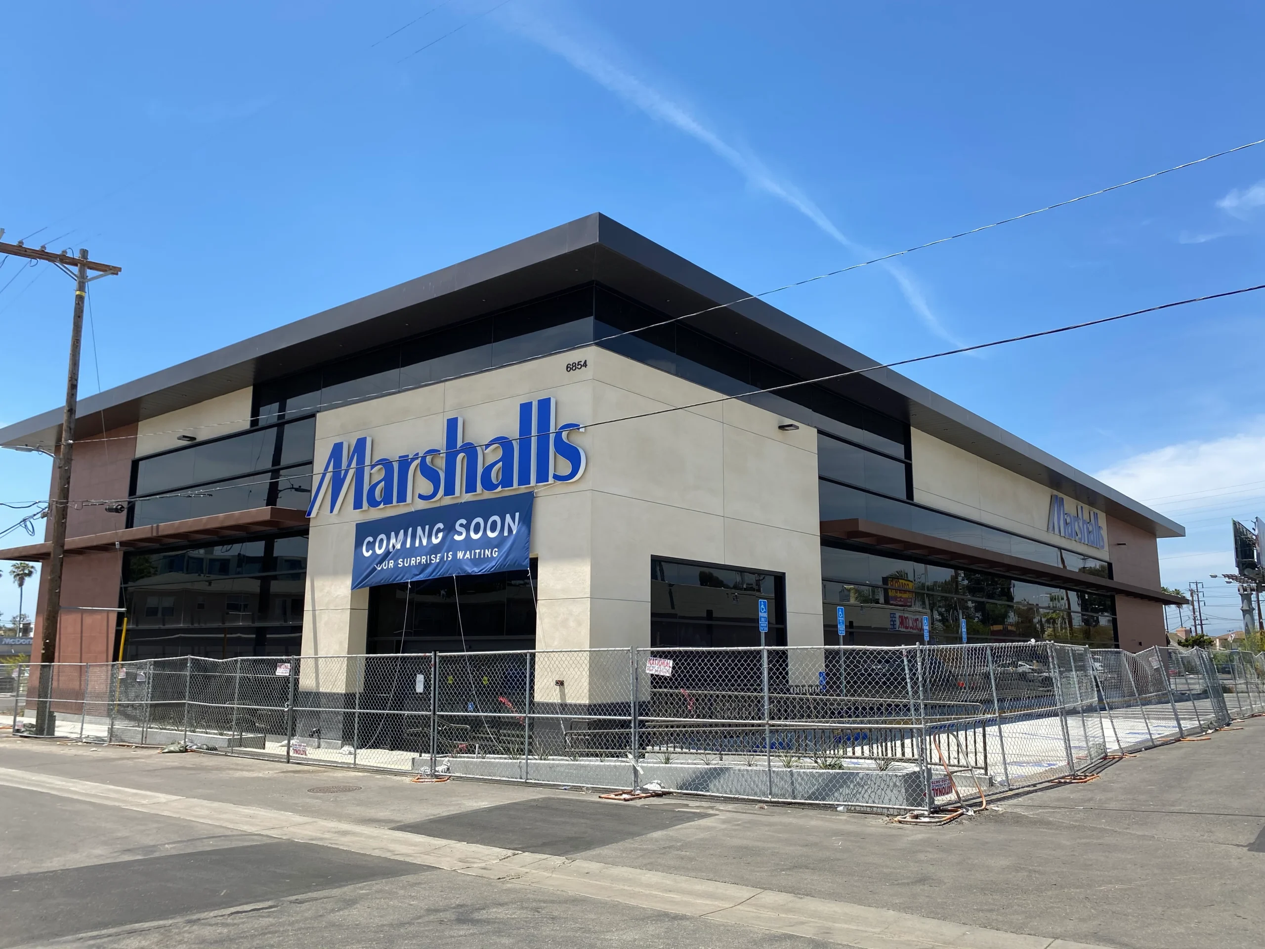 Building with a sign that reads "Marshalls Coming Soon" surrounded by a metal fence, a group of trees, utility poles
