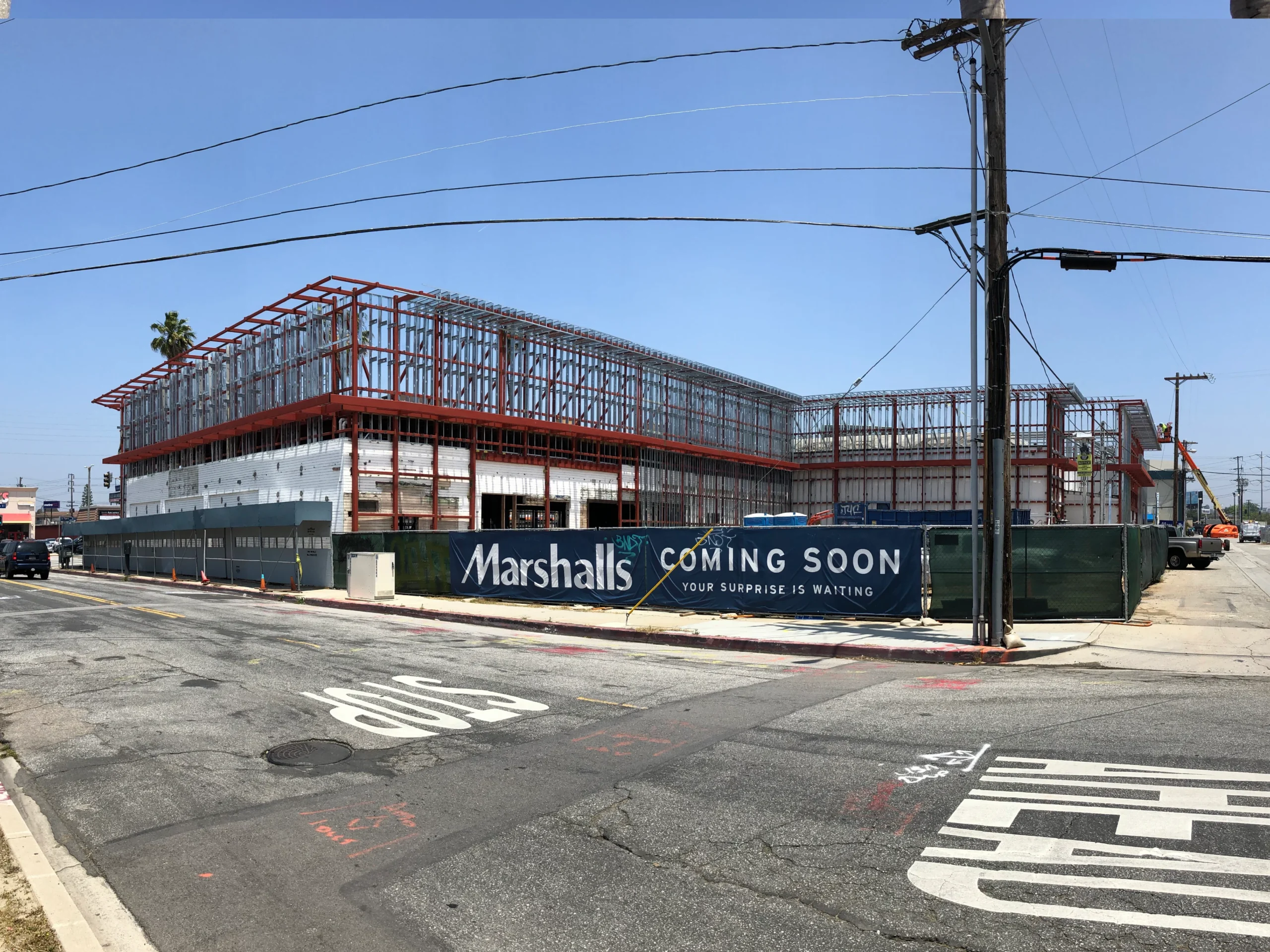 Large building under construction on the corner of a street, with vehicles driving by, a banner with text on it