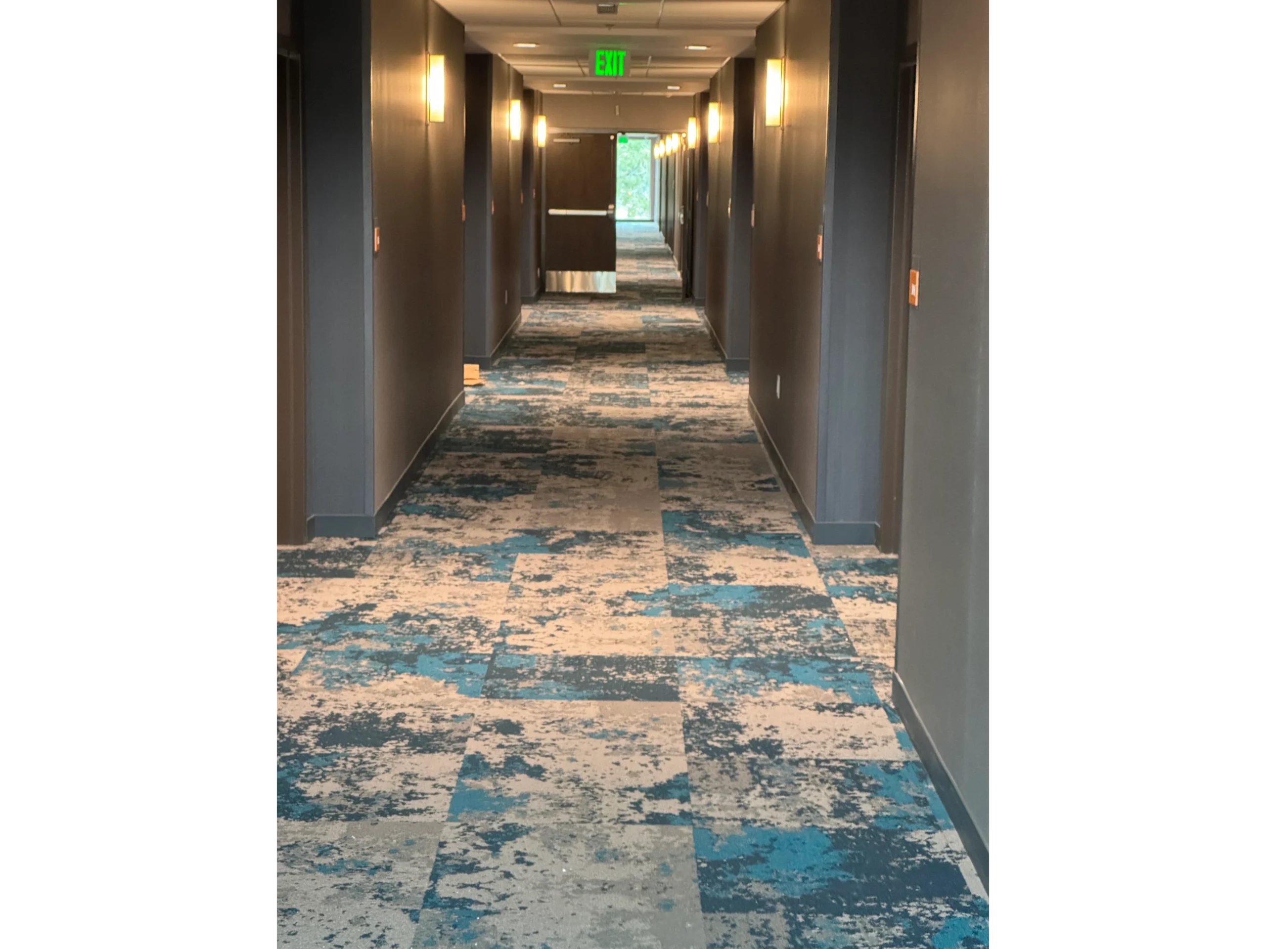 Long hallway with a blue and white patterned carpet, walls, lights, a door, and a ceiling with lights