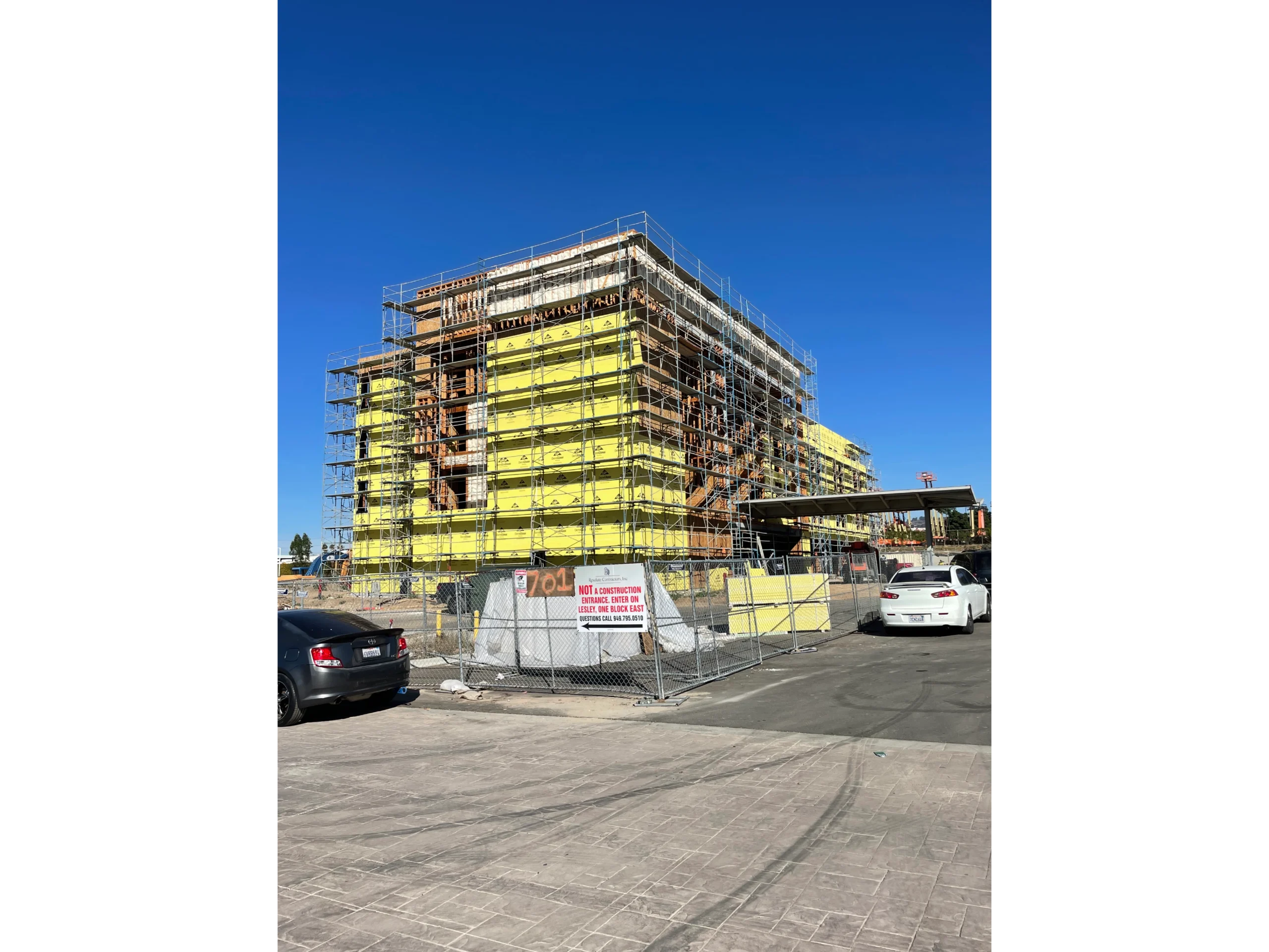 Large building under construction with scaffolding around it, surrounded by vehicles on the road, a fence, a board with text