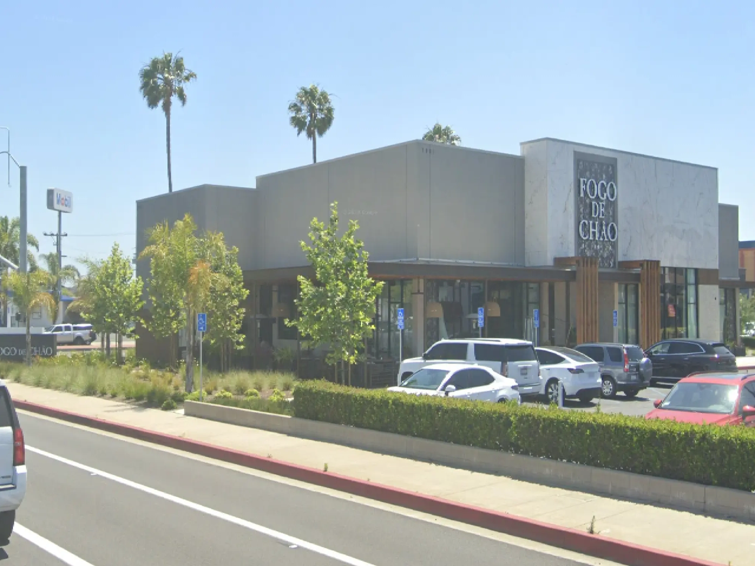 Building with cars parked in front of it, surrounded by trees, plants, poles, boards, and lights