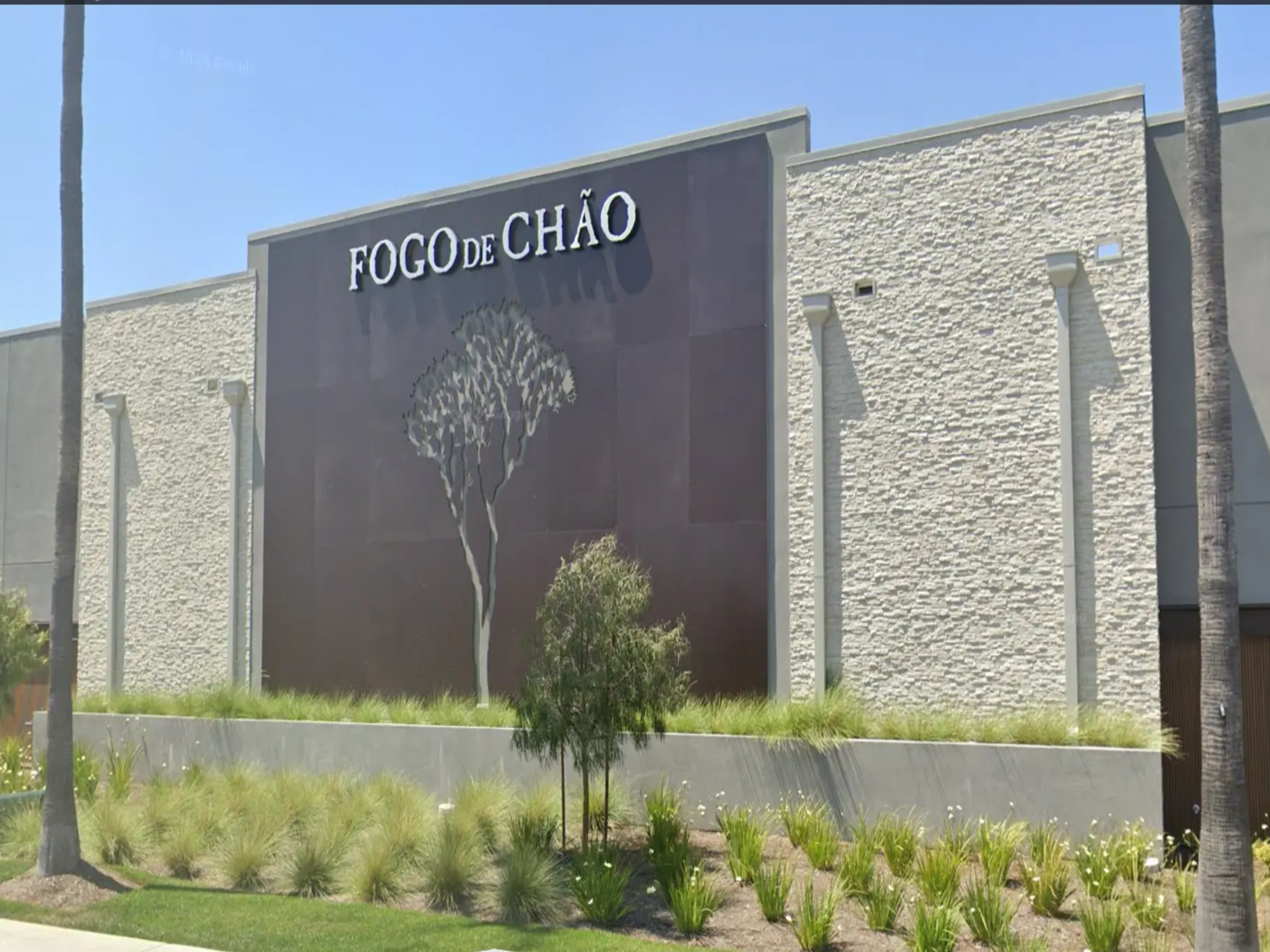 Building with a sign that reads "Fogo de Chão" surrounded by trees, plants, grass, and flowers
